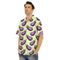 Man wearing a light green short-sleeve shirt patterned with purple eggplants and blue jeans, hands in pockets.